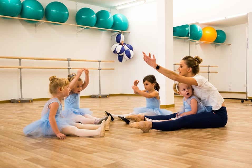 Tanzlehrerin sitzt auf dem Boden mit vier Vorschulmädchen in blauen Tutus in einem modernen Tanzraum.